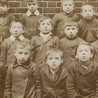 A sepia-toned photograph of a group of young boys at the Jewish Free School of London. According to the record, the boys are new immigrants from Russia who do not speak English. The boys are staring intently and directly at the camera. They are dressed similarly in collared, dark-colored jackets. Photo Credit: Photographer Unknown (1908). Source: Jewish Museum of London