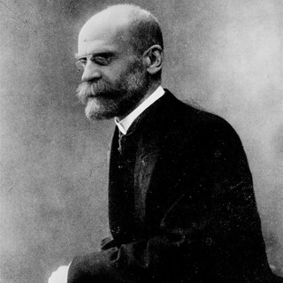 A grayscale photograph of Emile Durkheim in a dark jacket with a light shirt. He is seated facing sharply to the left, and has a stern look on his face. He is wearing round, thin glasses and he has a very full beard as well as moustache that are well-groomed and neat. He has a nearly bald head. No photo credit information. Source: commons.wikimedia.org
