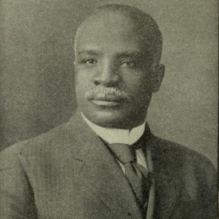 A sepia-toned photo portrait of Kelly Miller. He is wearing a suit and is sat perfectly upright. He has a white moustache and a serious, contemplative facial expression.