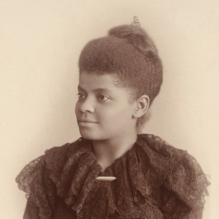A sepia-toned photographed portrait of Ida B. Wells. She is wearing a dark-colored dress with lacy frills around the collar. The dress has a gold clasp at the front. Her hair is nestled up into a full bun. Her head is turned to the left so she is looking away from the camera. She is smiling in a way that conveys self-confidence and mischief. Credit: Mary Garrity, 1983. Source: commons.wikimedia.org