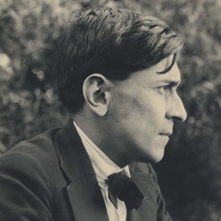 A sepia-toned photograph of José Carlos Mariátegui's side profile. He is wearing a suit with a bow tie. He has a serious and focused look on his face. There are trees in the background. Photographer: José Malanca (1928). Photo Source: Wikimedia Commons.