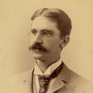 A sepia-toned portrait photograph of John Dewey. He is wearing a gray suit with a patterned tie. He has round-glasses and a thick mustache. His hair is dark and neatly combed along a middle part. He is looking blankly off to the left of the camera. Photo Credit: Gibson Photo (1980), Source: University of Michigan--Bentley Historical Library