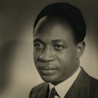 A sepia-toned portrait photograph of Kwame Nkrumah. He is wearing a dark colored suit with a white shirt and a tie. His hair is styled in a very round afro. He is looking slightly to the left of the camera into the distance with a rather straight face. Photo Credit: Undated, Unknown photographer. Source: The National Archives (United Kingdom)