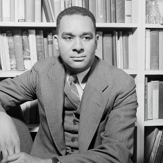 A grayscale photo portrait of a young Richard Wright sat in front of a wall of books in his study. He has hair that is cut short and a youthful face that appears very relaxed and confident.
