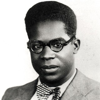 A black and white photo portrait of Aimé Césaire as a young man. His hair is cut into a somewhat squared afro. He is wearing round glasses, a sharp-looking suit, and a polka-dot bow-tie. He is looking intensely into the camera.