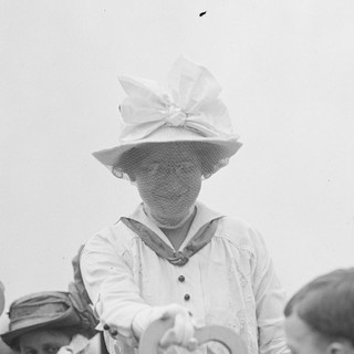 A gray-scale photo of Mary Ware Dennett at what seems to be a rally or gathering of some kind. She is wearing a light-colored dress with matching gloves. She is also wearing a marching hat that has a wide brim and a large bow on the front. She is wearing glasses. She seems to be smiling in kindly and waving at someone below her. Photographer: Harris and Ewing (1913), Source: Wikimedia Commons