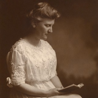 A sepia-toned photographed portrait of Mary White Covington. She is wearing a lacy dress and has her hair up in an intricate bun. She is looking down at a book, so the camera is only capturing the right side of her face. Source: By Charles J. Dampf / Adam Cuerden - This image is available from the United States Library of Congress's Prints and Photographs divisionunder the digital ID ppmsca.23826. Public Domain, https://commons.wikimedia.org/w/index.php?curid=139744190