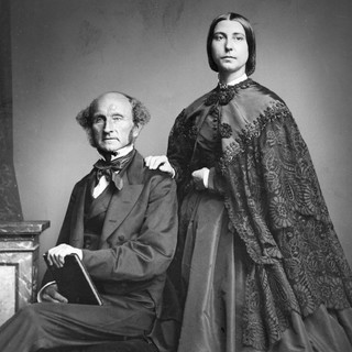 A photo portrait of John Stuart Mill and Harriet Taylor Mill. John is seated on a chair on the left-side of the photo and he is holding a book. Harriet is standing on the left side with her hand rested on his shoulder. They are both looking directly into the camera with serious and stoic facial expressions. John is wearing a suit and Harriet is wearing a long dress with a cloak. Year: 1869. Photographer unknown. Source: commons.wikimedia.org
