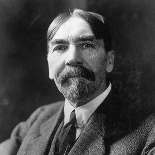 A black-and-white portrait photograph of Thorstein Veblen. He is wearing a tweed jacket, a white shirt, and a tie. He has a bushy moustache and bit of beard hair on his chin. His hair is smooth, dark, and parted down the middle. He is looking directly at the camera. He looked tired but determined. Photo Credit: photographer and year unknown. Source: commons.wikimedia.org