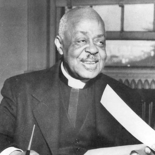 a black and white photo of Dr. Wright holding a piece of paper and smiling. He is wearing a black suit with the typical white pastor neckwear piece.