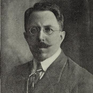 A sepia-toned photo portrait of Luis Cabrera wearing a suit and round-shaped glasses. He has a neatly-groomed moustache that points to the sides and thick black, slicked-back hair.