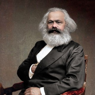 A colorized photograph portrait of Karl Marx. He is sitting rigidly upright with his hand inside his suit jacket in an upholstered wooden chair. He has a large gray beard and somewhat long hair to match. He looks very serious. / photo by: John Jabez Edwin Mayall. Colored by: Olga Shirnina. Year: 1875. https://commons.wikimedia.org/wiki/File:Karl_Marx,_1875.jpg#/media/File:Karl_Marx,_1875.jpg