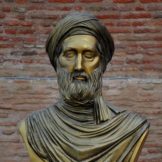 A bronze bust statue of Ibn Khaldun. The statue has a full, dark-colored beard. The bust depicts Khaldun in a neatly-wrapped turban and with scarves draped and wrapped over his shoulders. The bust depicts Khaldun with a serious facial expression. The real statue is located at the entrance of the Kasbah of Bejaia, Algeria. Photo taken by Reda Kerbush.