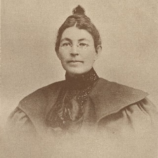 A sepia-toned photo portrait of a college-aged Mary Roberts Coolidge (maiden name Smith). Her dress has a high neck and her hair is in a small and high bun on her head. She is wearing small, thin, and round glasses.