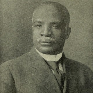 A sepia-toned photo portrait of Kelly Miller. He is wearing a suit and is sat perfectly upright. He has a white moustache and a serious, contemplative facial expression.