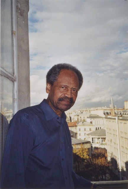 Photo of James A. Emanuel with a blue shirt standing at an open window in Paris.