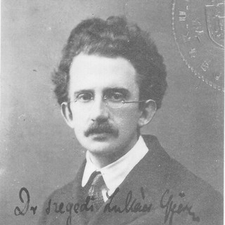 A grayscale photo of Georg Lukacs looking directly at the camera with a straight face. He is wearing round glasses, a dark coat, a light shirt, and a tie. He has a narrow moustache and a full head of curly hair. At the bottom of the photo there is some text written in cursive that I cannot understand. Credit: unknown, 1917. Source: commons.wikimedia.org