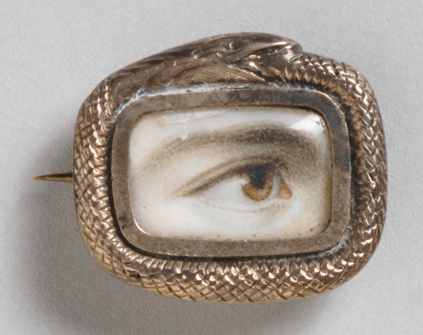 A miniature watercolor portrait of a single human eye, painted on ivory and set in a small oval gold brooch; the eye and brow gaze outward from within a thin gold frame.