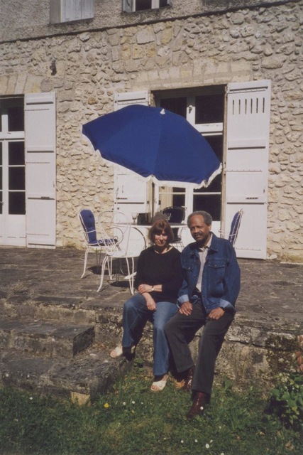 Marie-France Plassard on the left and James A. Emanuel on the right. Dressed causally, on the raised porch of Marie-France's Le Barry home in Condom, France. The home is tan with a rock structure and French styled-shutters.