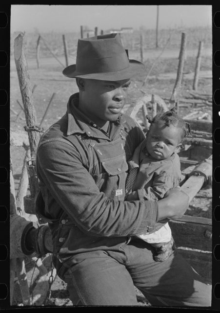 Depicts a Black man holding a baby. The man has on a hat and overalls. They appear to be sitting on some type of farm equipment. The photo is in black and white.