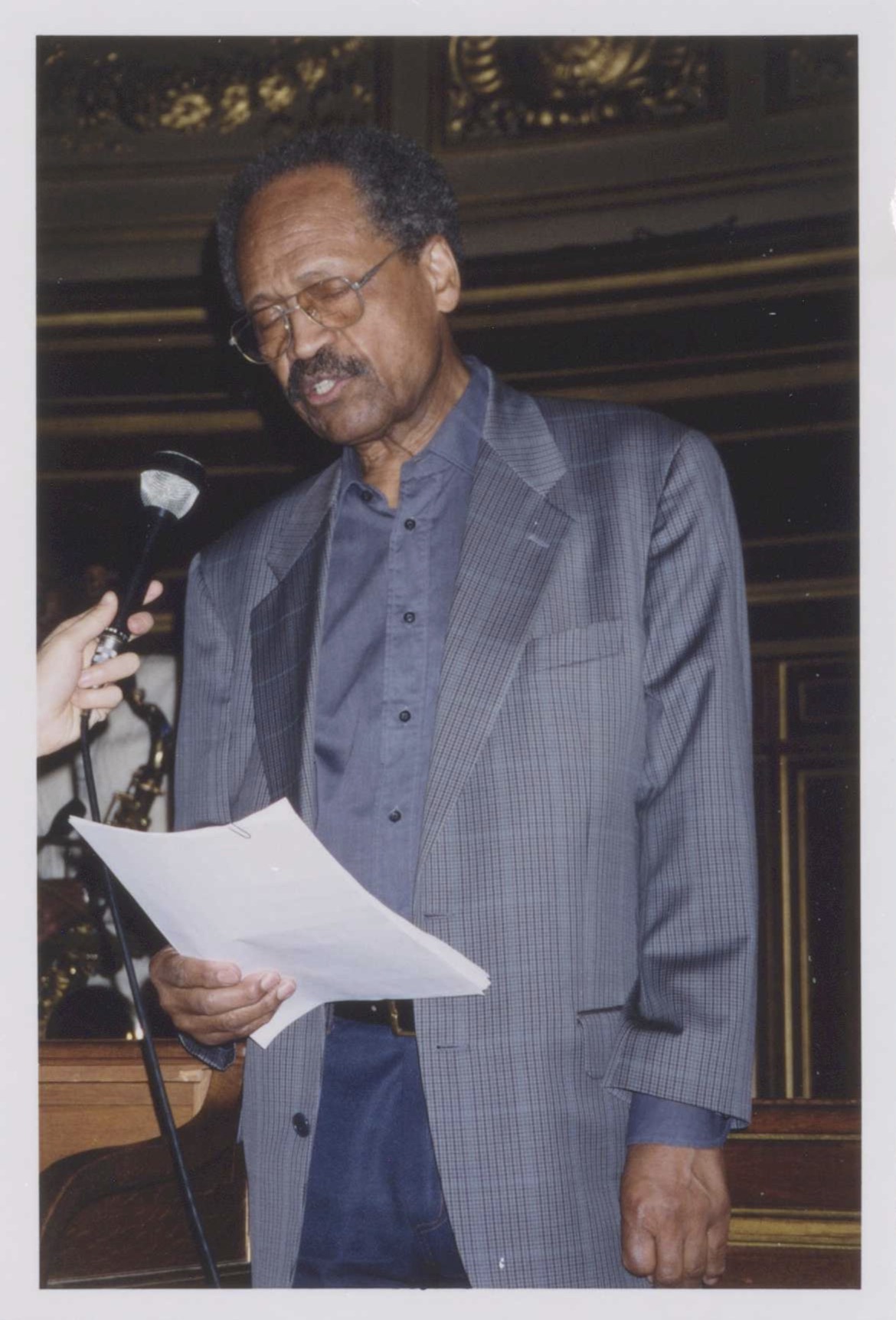 James A. Emanuel dressing in blue. A microphone is held to his mouth by an unknown person. He is wearing glasses, and his eyes are closed. He is holding sheets of paper.