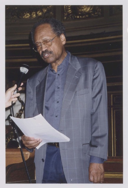 James A. Emanuel dressing in blue. A microphone is held to his mouth by an unknown person. He is wearing glasses, and his eyes are closed. He is holding sheets of paper.
