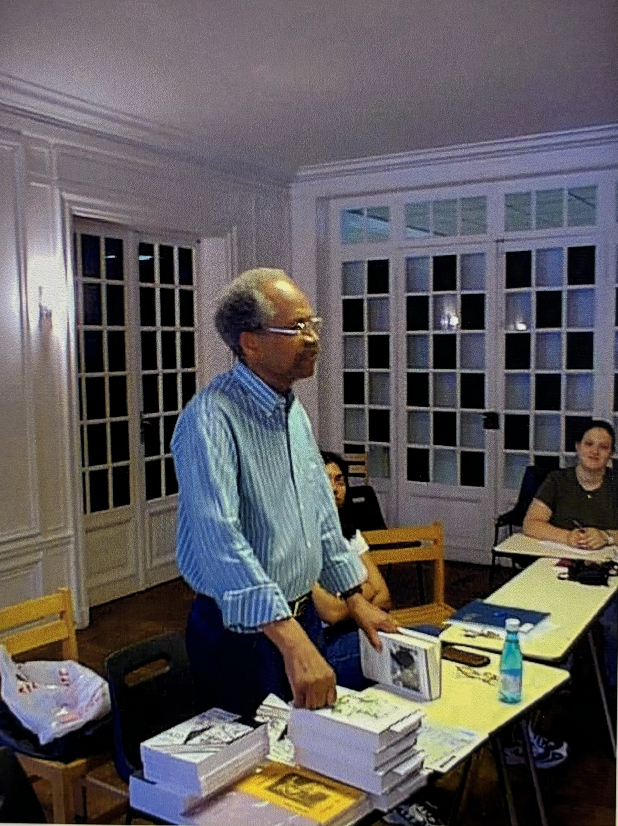 James A. Emanuel standing over a desk with one of his books in his hand. He has a stack of books near him, which look to be 'Jazz from the Haiku King' and 'Whole Grain: Collected Poems 1958 to 1989.' Two students are looking at him.