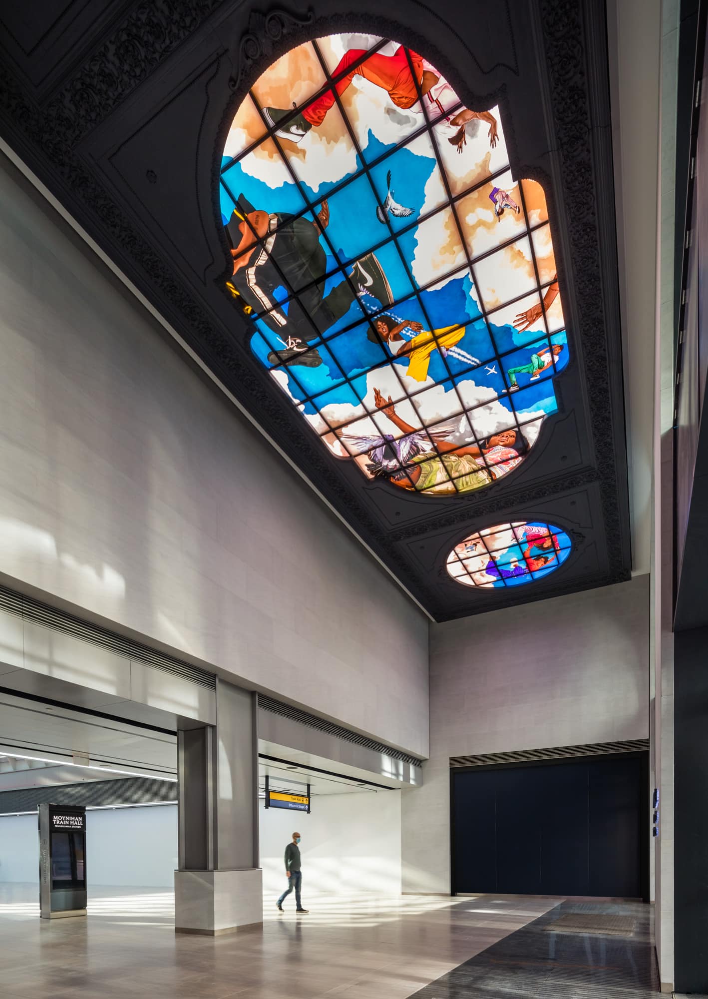 Angled view of 2 our of the 3 stained glass windows on the ceiling of the Moynihan train station, featuring Black youth in various dancing and running poses, with a train commuter in the foreground.