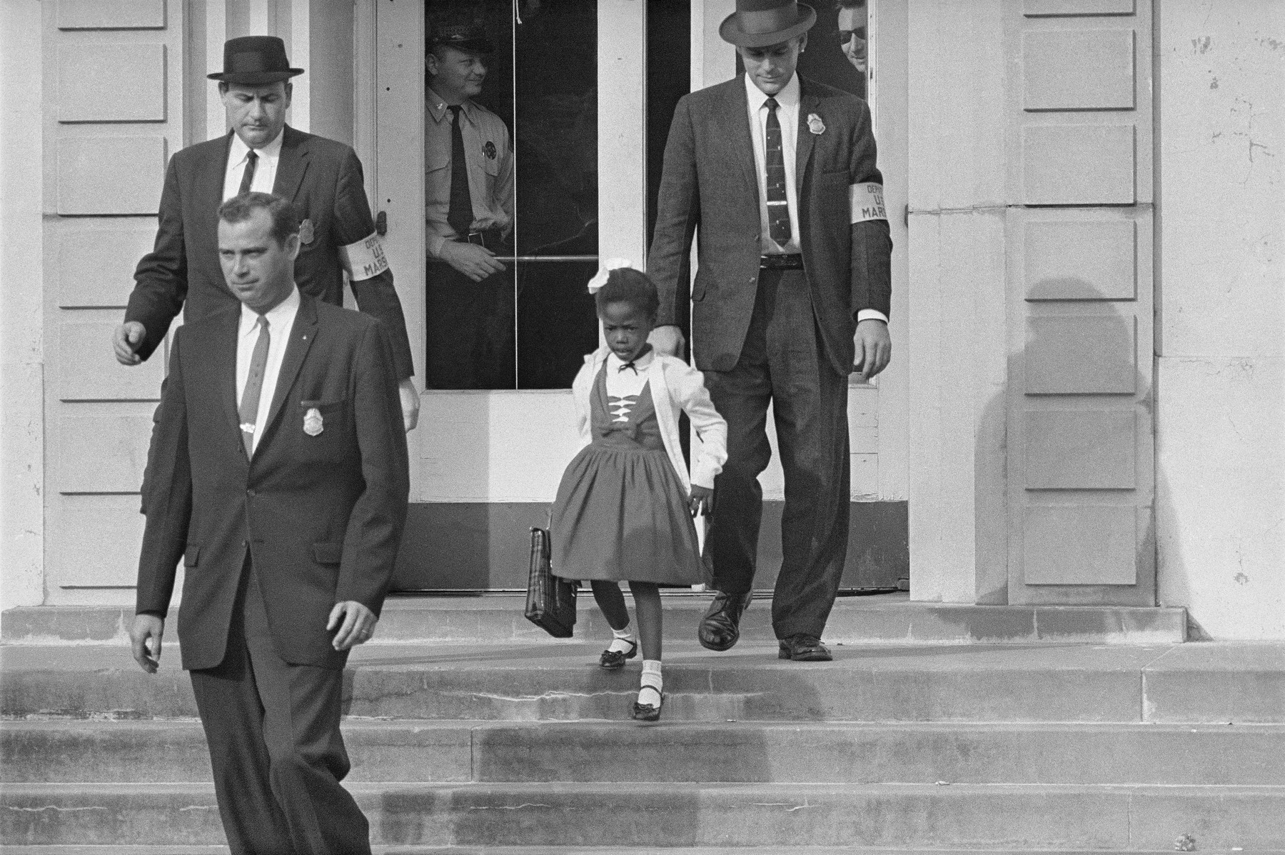 A young Black girl in a white dress and white hair ribbon walks down the steps of a school building, escorted on either side by four men in suits and armbands. She carries a notebook and ruler and looks ahead; the men's faces are above the frame.