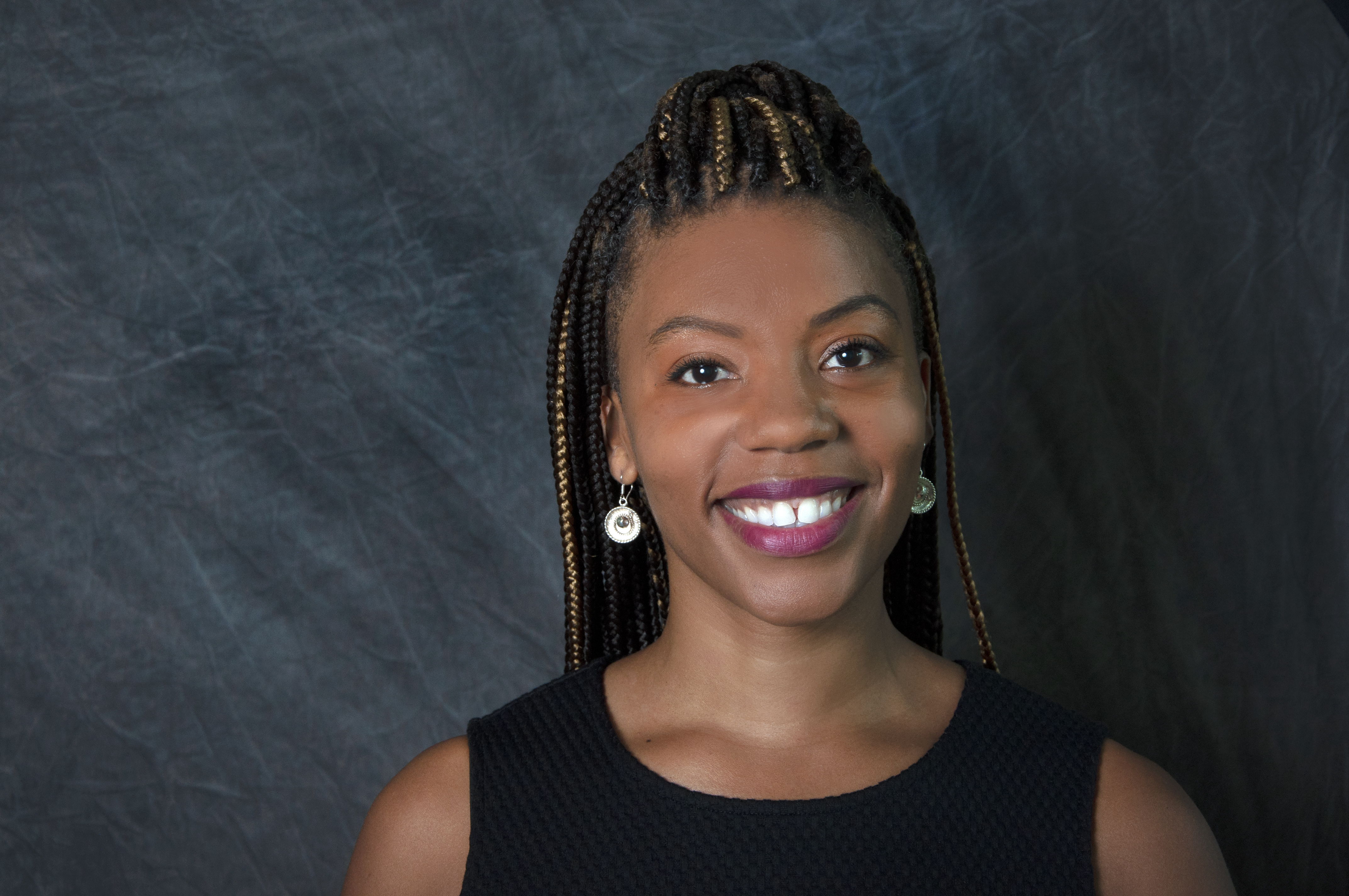 Photograph of Tyechia Thompson, a Black woman smiling with brown and blond box braids, wearing a black top without sleeves, and silver earrings. She is standing against a bluish grey background.