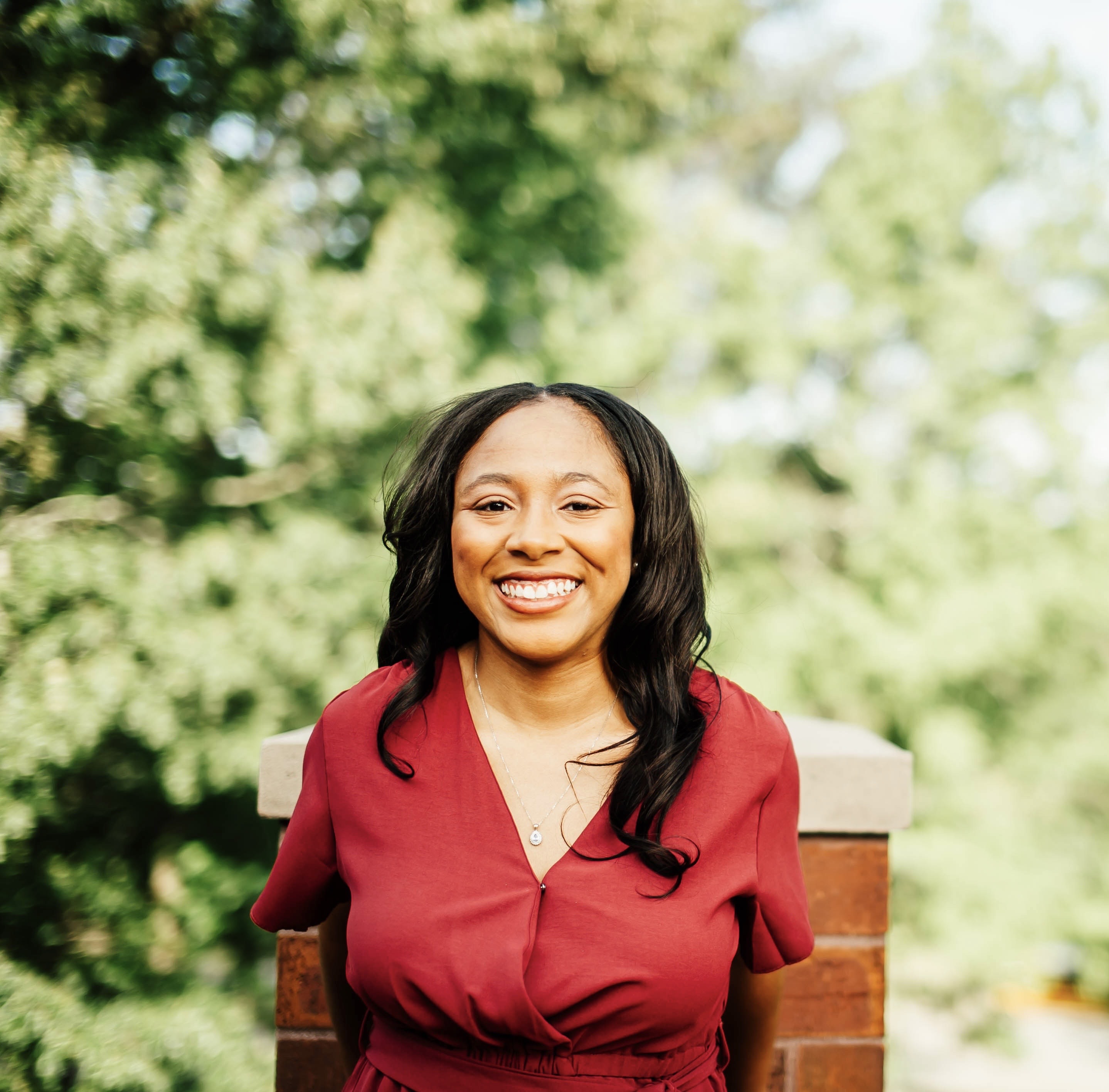 A young Black woman wearing a burgundy blouse smiling outdoors. 