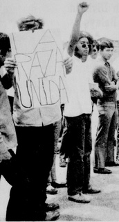A student protester raises a fist while another holds a sign reading "La Raza Unida"