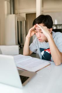 a student looking at a laptop and textbook with his hands on his temples looking stressed, exasperated, and confused