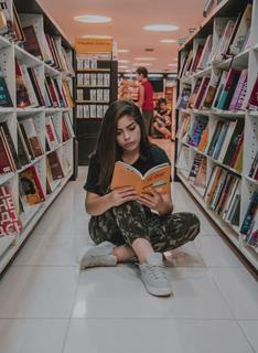 This is a photo of a woman reading a book while sitting on the tiled floor between two library bookshelves.