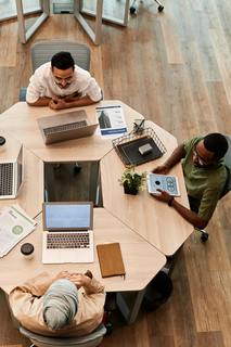 A top-down photograph of 3 students at an octagonal table talking with eachother in a friendly way. there are laptops, papers, phones, and other items all over the table. Photo by Edmond Dantès (pexels.com)