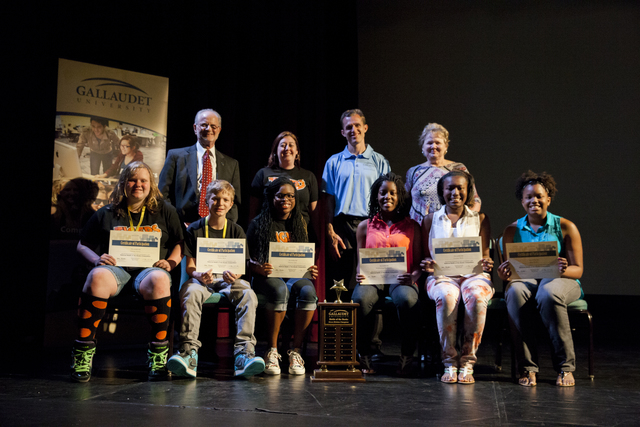 Six young people sit in chairs on a dark stage, each holding a paper certificate and smiling. From the left, there is a young white blonde girl with a black t-shirt with orange lettering and denim shorts. Her socks are black with large orange spots. Her shoes have neon green laces. Next to her is a small white blonde haired boy with a black t-shirt, khaki pants, and light blue sneakers. Next to him is a Black girl also wearing a black shirt with orange lettering and denim capris with the cuffs rolled up. She has black rimmed glasses and long braided hair. A large trophy with a star on top sits on the ground between her and the next young girl. She is also Black and is wearing a pink sleeveless top and dark pants. To her left is another girl, Black, in a white sleeveless top, pink and white floral pants, and white sandals. Her hair is pulled back with a headband. The final young person is a Black girl in a blue sleeveless top, dark pants, and sandals. Her hair is also pulled back with a headband. Behind the seated children, stand four white adults. The first on the left is an older man with white hair and a white beard and mustache. He's wearing a dark suit with a red patterned tie. Behind him is a large poster with the words "Gallaudet University" and the swoop logo.  Next to him is a short woman with dark hair, wearing a black t-shirt with orange lettering. Next is a tall dark-haired young man, clean shaven, with a light blue shirt. And finally stands a blonde woman wearing a black and red pattern shirt.
