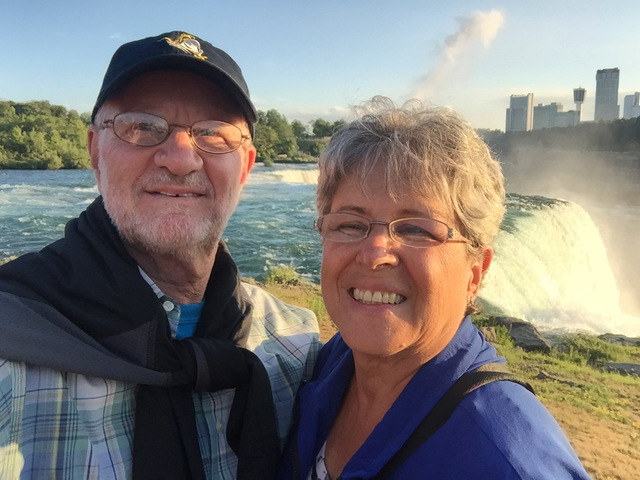 An older man and woman stand in front of water cascading over cliffs. The man has a white beard and glasses and is wearing a dark blue baseball cap with a dark coat tied around his neck. He has on a blue, white, and yellow plaid buttoned-up shirt with a light blue t-shirt underneath. The woman has wire-rimmed glasses, short salt and pepper hair and is wearing a blue jacket. Behind her shoulder are large buildings in the distance.