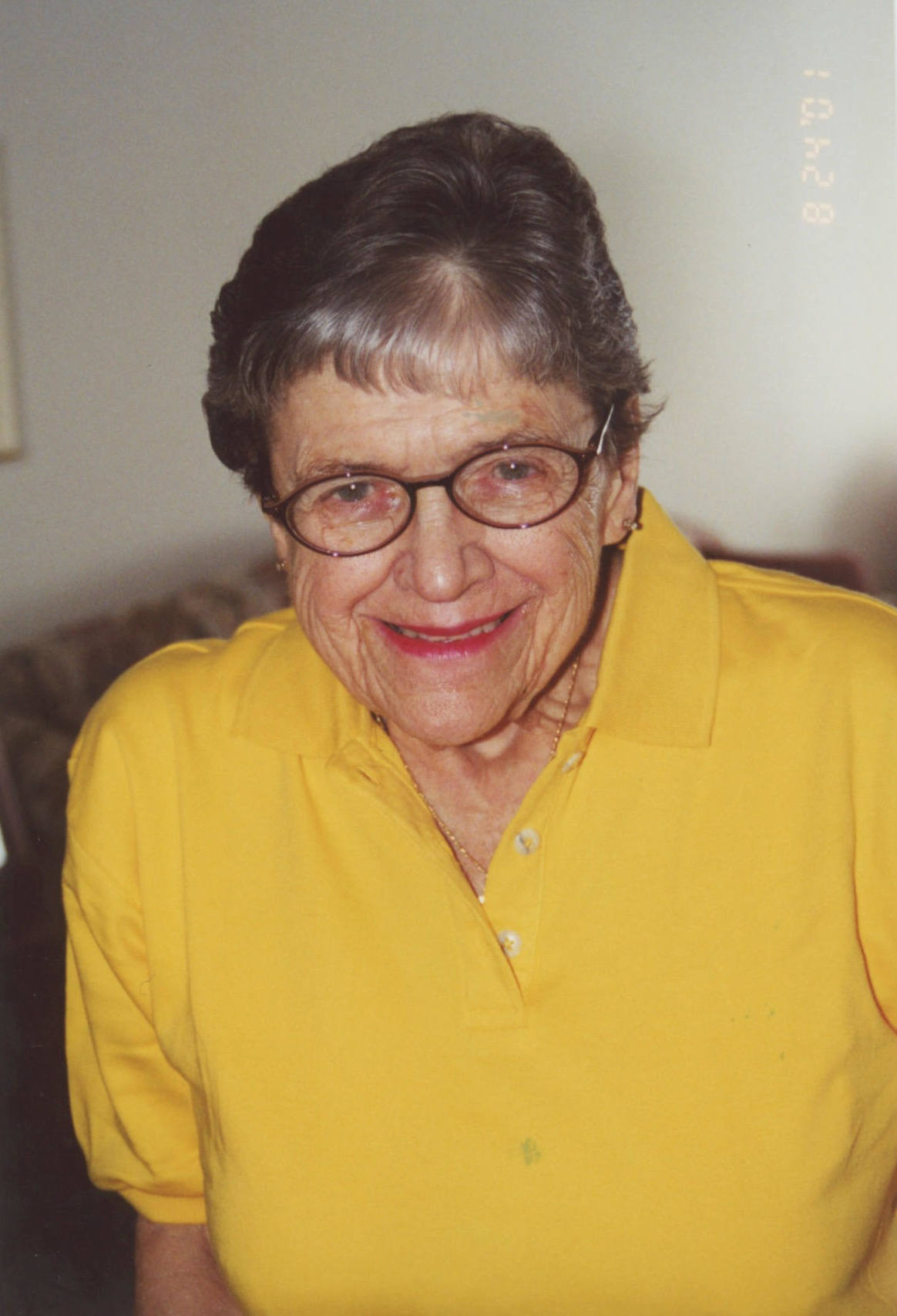 An elderly woman in a bright yellow polo shirt smiles at the camera. She has short dark gray hair and is wearing red lipstick. She has oval glasses with dark brown frames, stud earrings, and a gold necklace.