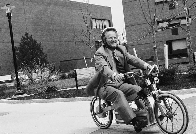 Black-and-white photo of a middle-aged smiling white man in a suit with a helmet on his head riding a low slung bicycle on a brick walkway. Beneath the seat of the bicycle is a motor.