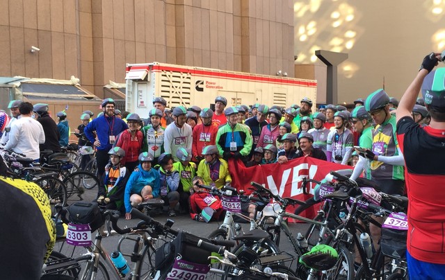 A large group of predominantly white people are standing for a photo on a street in New York City. Most are wearing gray bike helmets with a neon green streak down the center. In the foreground are parked bicycles with purple and white tags mounted between their handlebars. The tags have numbers printed in black..
