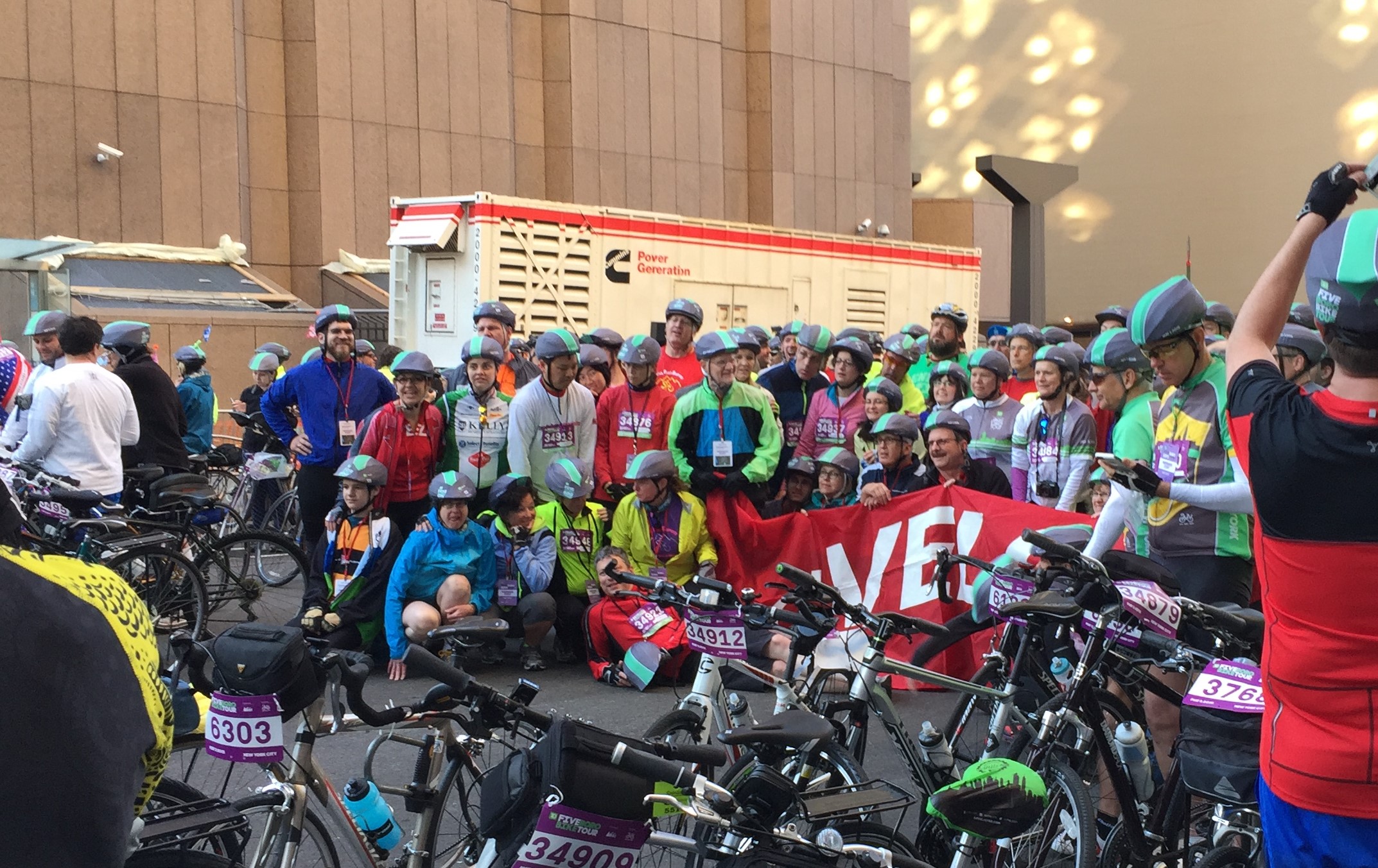 A large group of predominantly white people are standing for a photo on a street in New York City. Most are wearing gray bike helmets with a neon green streak down the center. In the foreground are parked bicycles with purple and white tags mounted between their handlebars. The tags have numbers printed in black..