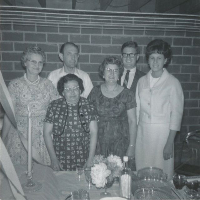 Black and white photo of a group of white people standing in front of a brick wall. There are two elderly women on the left side, both wearing dresses. To the right of them stands an older white man and woman. Next to them are a young man wearing black glasses and a striped tie and a young woman in a light colored dress. In front of them is a table with a flower arrangement, a punch bowl, and glasses.