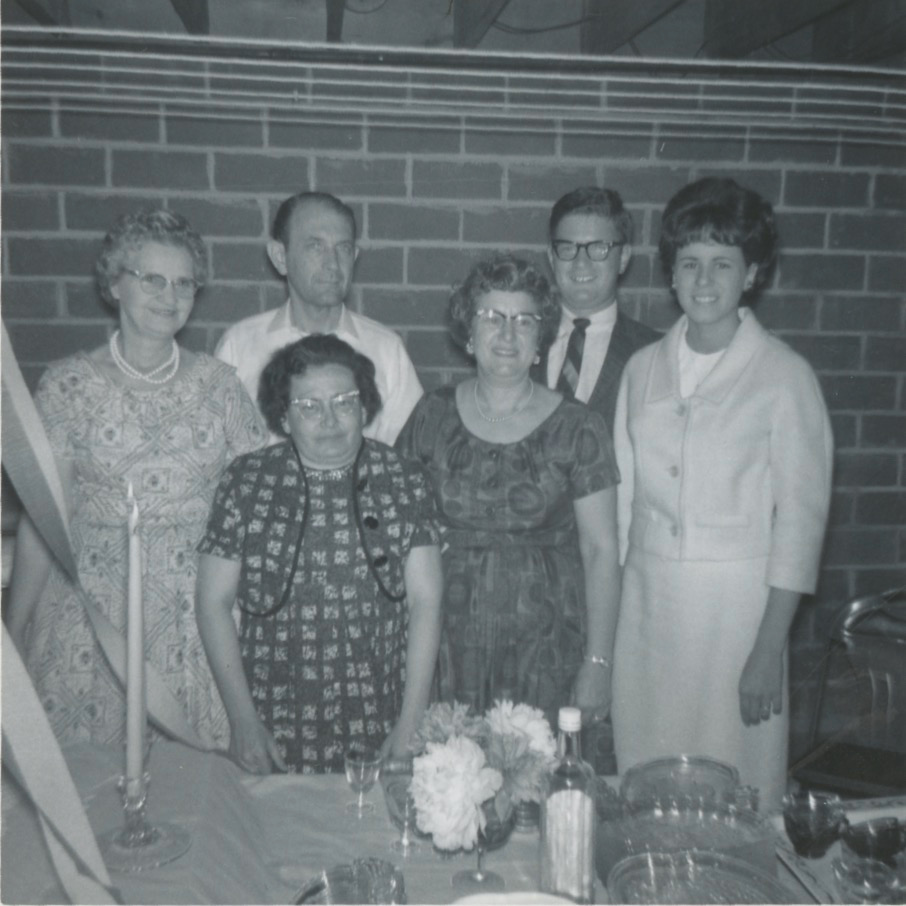 Black and white photo of a group of white people standing in front of a brick wall. There are two elderly women on the left side, both wearing dresses. To the right of them stands an older white man and woman. Next to them are a young man wearing black glasses and a striped tie and a young woman in a light colored dress. In front of them is a table with a flower arrangement, a punch bowl, and glasses.