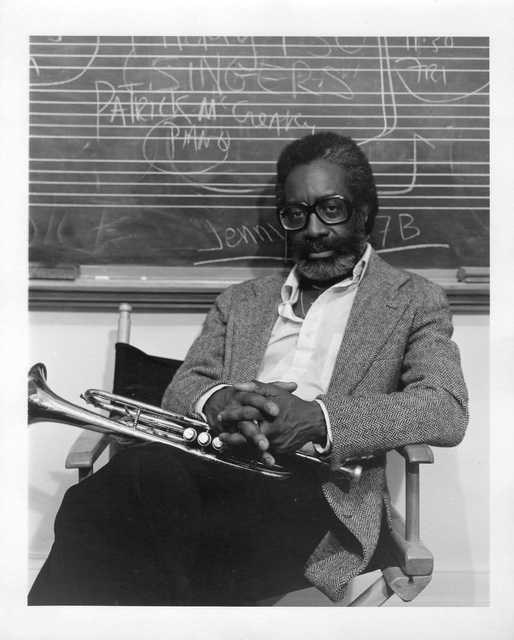 Bill Dixon, seated, holding a trumpet in his lap, in front of a chalkboard with writing on it.