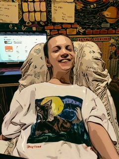 A boy with open, smiling, and playful face is looking at the camera; he is sitting in a chair dressed in a white t-shirt with what seems like a naïve picture of wolf and full moon. Behind him, there is a on open laptop gleaming with light and rows of school-related rules and a poster of Solar system hanging on the wall.