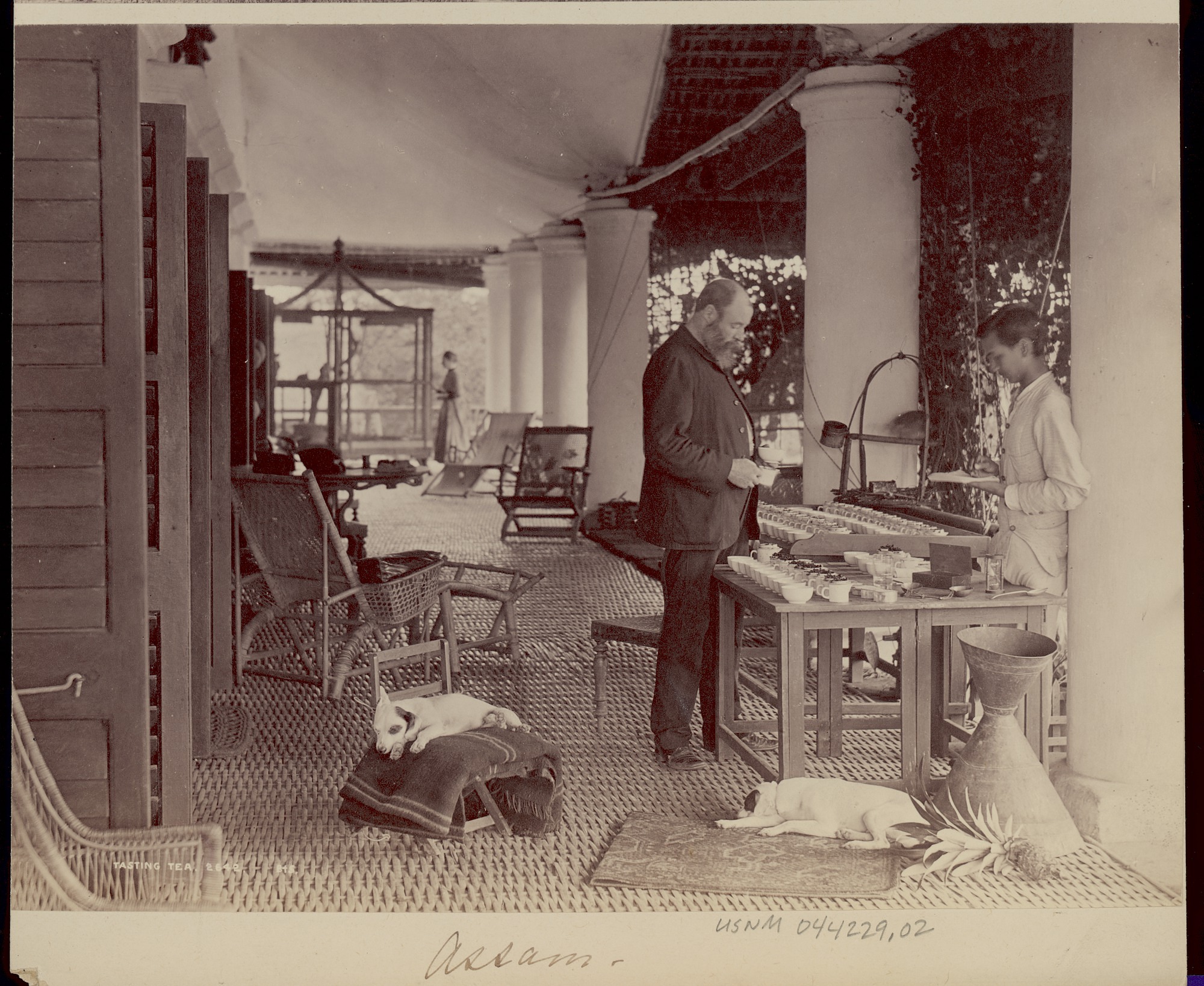 On an outdoor veranda a white man, likely British, tastes different teas from an Assam man.