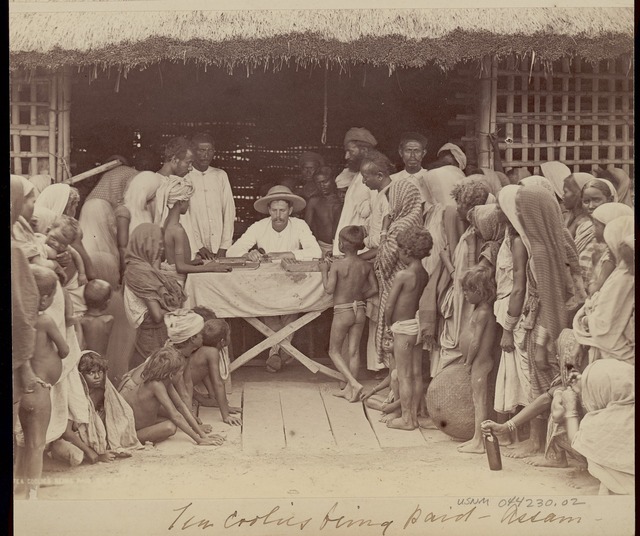 A white man sits center in the photo while on both sides of him there are lines of men, women, and children waiting to be paid