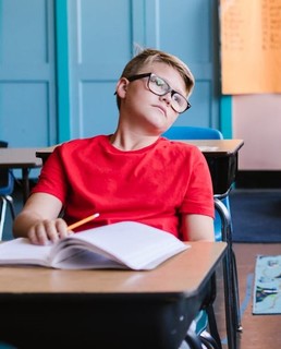 A boy in a red shirt, sitting at a desk, looking off into space.