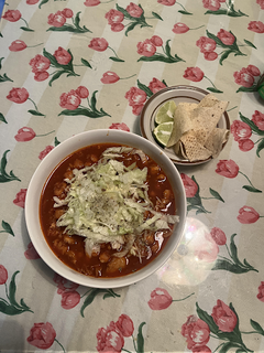 Red broth-based soup in a white bowl on a flowered tablecloth, with a plate of tortilla chips and sliced limes beside