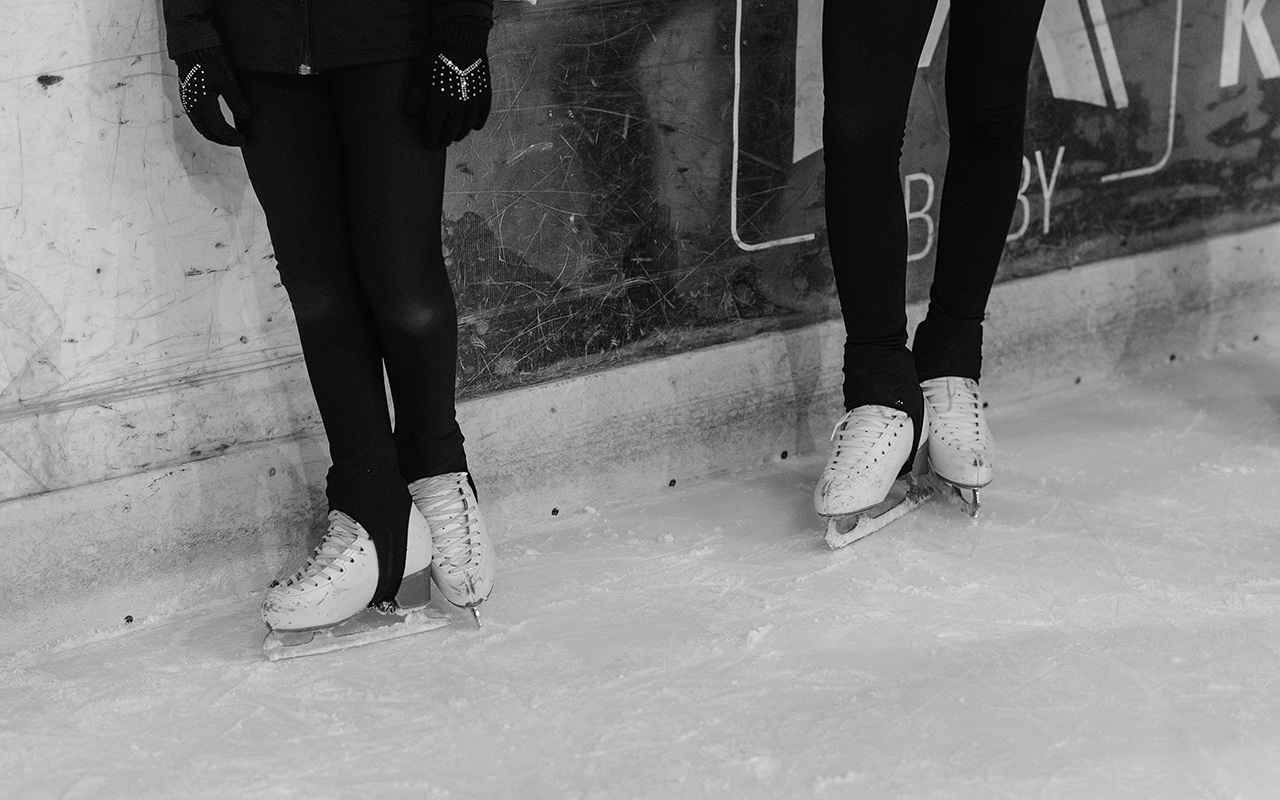 Two people in black leggings in an ice rink wearing ice skates.
